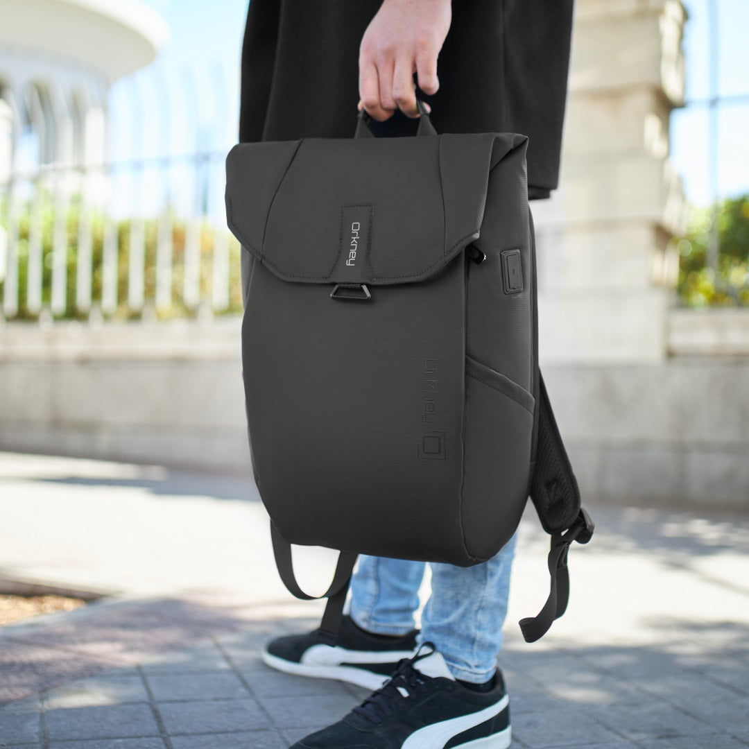 Imagen de una mochila Orkney en color negro, colocada sobre un fondo urbano. La mochila presenta un diseño moderno y funcional, con compartimentos ocultos.